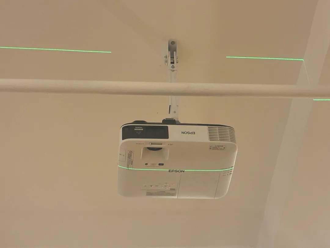 A white projector hanging from a ceiling arm under a pipe, with two green laser lines across the ceiling.