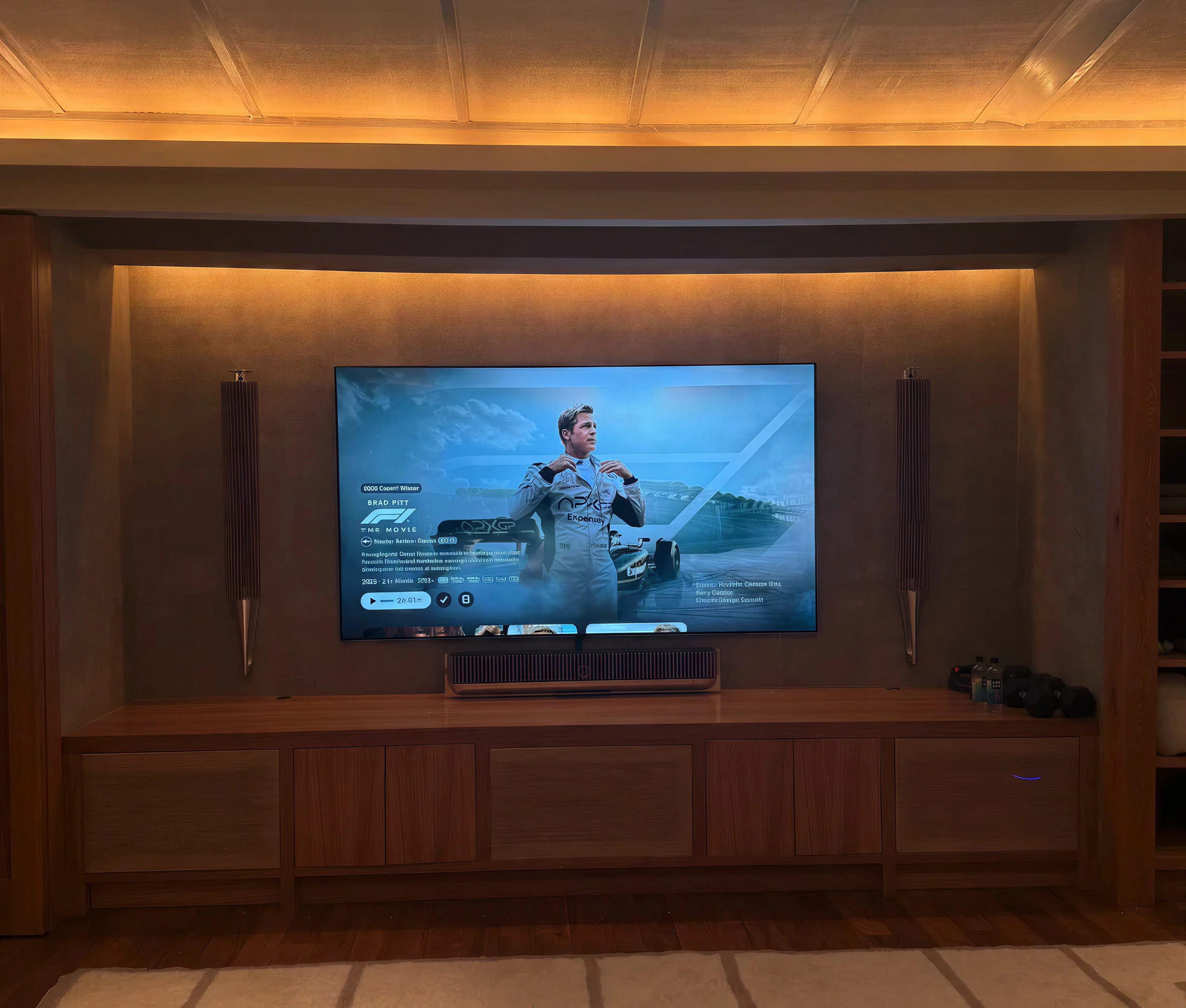 A large TV in a cove-lit recess between walnut built-ins, with Bang & Olufsen speakers and a soundbar on a built-in console, in a home theatre